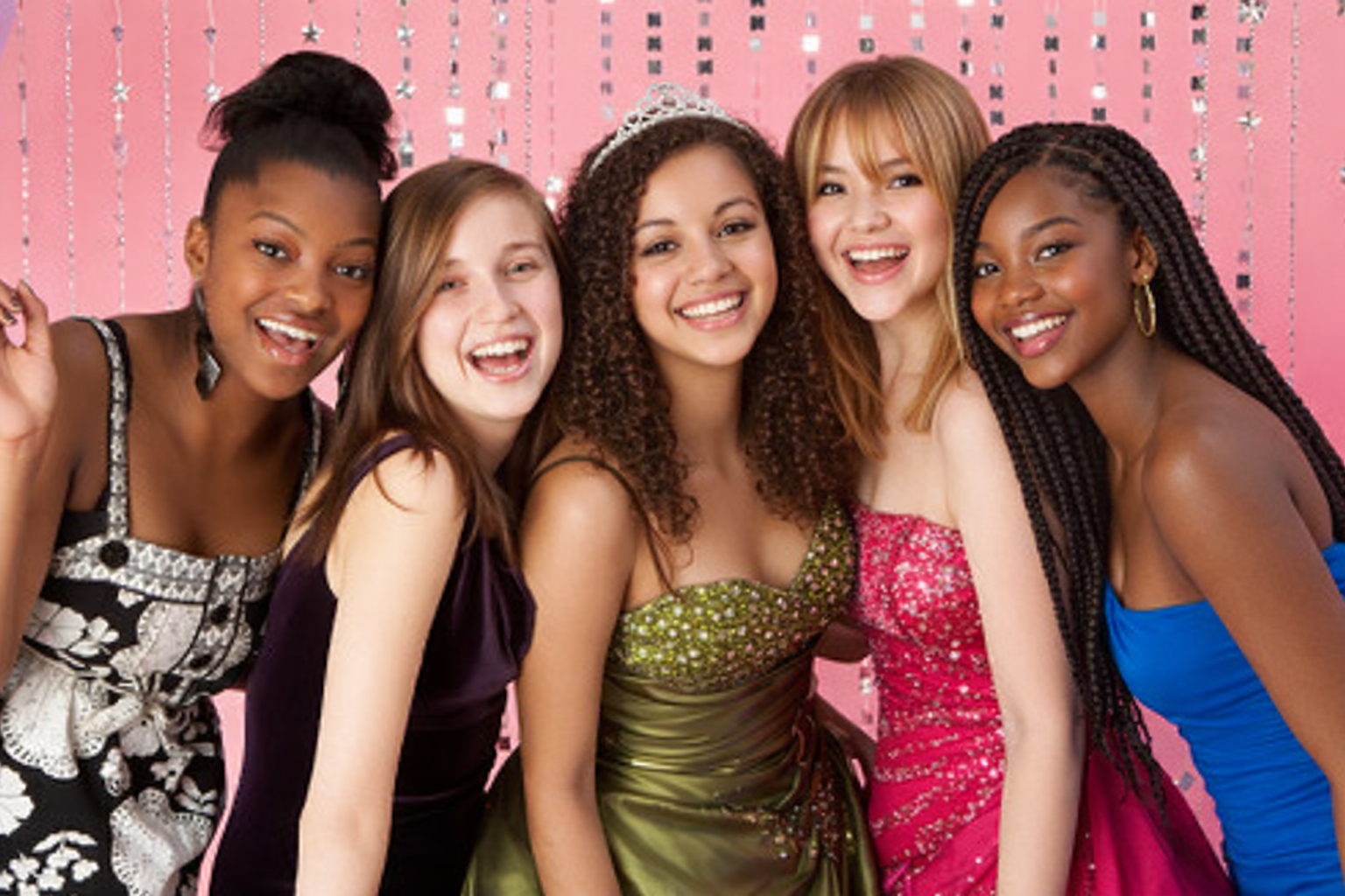 Group of diverse young women smiling together in elegant formal dresses against a pink, sparkling backdrop